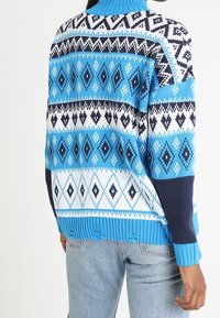 Blau-weißer gemusterter Pullover mit hohem Kragen, der Rautenmuster und einen gerippten Saum aufweist, gestylt mit langen Ärmeln und einem lockeren Schnitt.