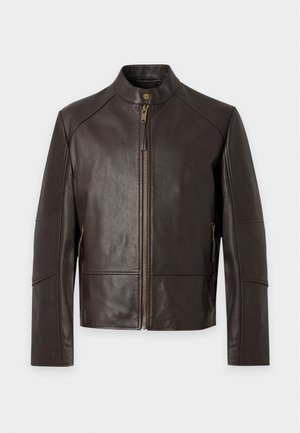 Homme debout portant une veste en cuir marron, un t-shirt bleu, un jean foncé, des baskets beiges et une ceinture noire sur un fond blanc uni.