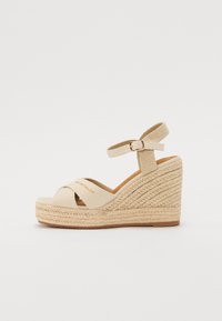 Beige kile sandal med en flette jute mellomsåle og kryssremdesign. Ankelrem med spent lukking. "Tom Tailor"-logo synlig.