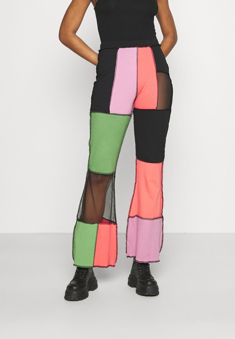 Femme portant un pantalon patchwork taille haute avec des panneaux en mesh verts, roses, corail et noirs, associé à des bottines à plateforme noires et un haut noir.