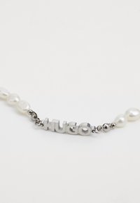 Colier cu lant de ton argintiu, avand un text lucios "HUGO", intercalat cu perle albe neregulate si accente metalice.