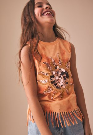 Ung pige, der griner og har en ærmeløs orange top med solformet palietdesign og frynsekant, kombineret med blå jeans.