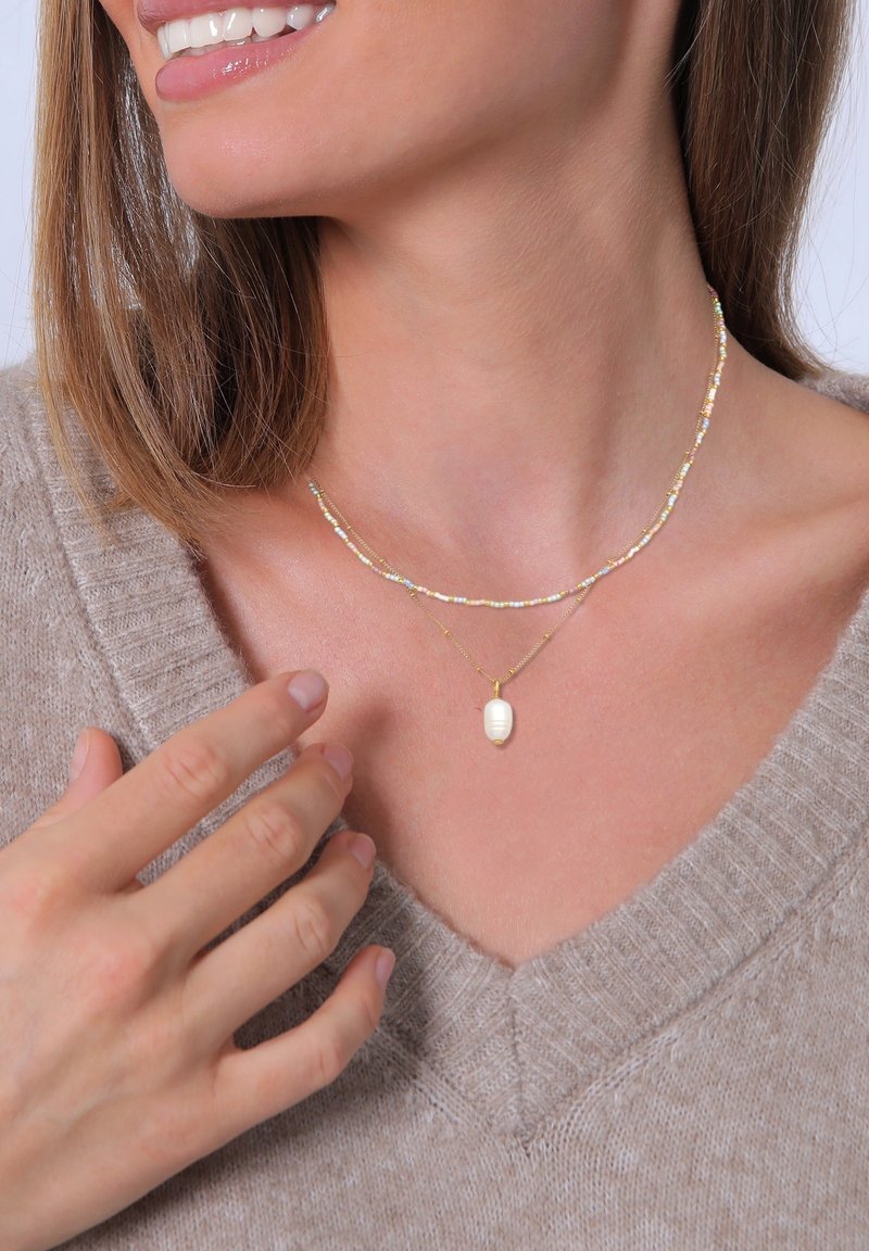 Femme portant deux colliers, l'un avec des petites perles et une chaîne en or avec un pendentif en perle blanche, souriante et touchant son pull.