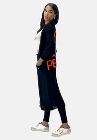 Lång svart cardigan med färggmönster, stickat tyg, öppen front, sidofickor, bärs över en krämfärgad topp och svarta leggings, kombinerad med vita sneakers.