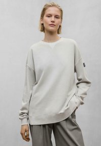 Sweatshirt gris clair avec un col rond, des épaules tombantes et des poignets côtelés. Texture douce ; coupe ample et décontractée. Design minimaliste sans motifs.