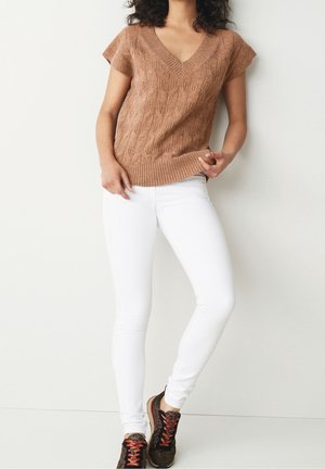 Femme portant un haut en maille marron à manches courtes, un pantalon slim blanc et des baskets marron avec des lacets rouges, debout contre un mur clair uni.
