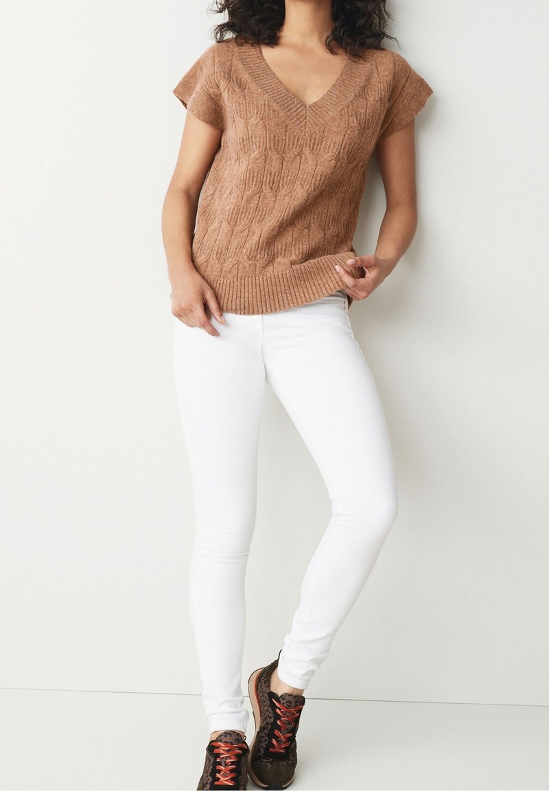 Femme portant un haut en maille marron à manches courtes, un pantalon slim blanc et des baskets marron avec des lacets rouges, debout contre un mur clair uni.