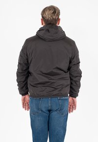 Giacca nera imponente con cappuccio, dotata di polsini e vita elasticizzati. Indossata sopra dei jeans blu, vista da dietro su uno sfondo semplice.