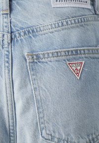 Lyseblå denimjeans med et trekantet rødt og hvitt "GUESS"-logo på baklommen, med synlige sting og glatt tekstur.