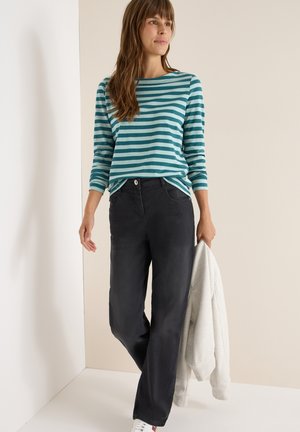 Kvinde iført en blåt og lyseblåt stribet langærmet skjorte, sorte bukser, hvide sneakers, der holder en lys grå jakke, stående mod en beige baggrund.