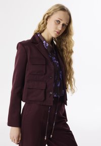 Burgunderfarbener, taillierter Blazer mit vier vorderen Taschen, zwei Knöpfen und schmalem Revers, kombiniert mit einer lila Blumenbluse. Glatte Textur, passgenauer Schnitt.