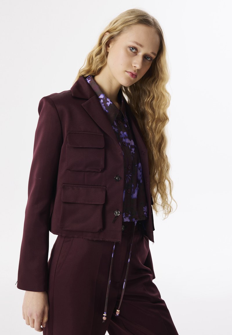 Burgunderfarbener, taillierter Blazer mit vier vorderen Taschen, zwei Knöpfen und schmalem Revers, kombiniert mit einer lila Blumenbluse. Glatte Textur, passgenauer Schnitt.