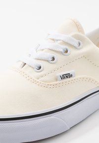 Baskets Vans en toile beige avec lacets blancs, œillets métalliques, coutures visibles et semelle en caoutchouc blanche avec une bande noire.