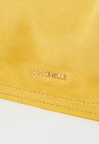 Matériau en daim jaune avec une texture lisse, présentant un logo en relief au design « COCCINELLE » et de fines coutures le long des bords.