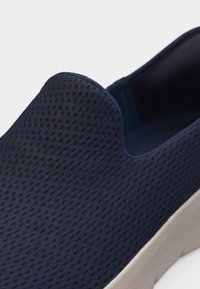 Παπούτσι σε σκούρο μπλε χρώμα με εύκαμπτο και διαπνέον πάνω μέρος από δίχτυ και λευκή σόλα, με κοντινή λήψη των μπροστινών και πλευρικών λεπτομερειών.