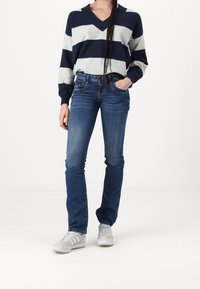 Pull oversized rayé bleu marine et gris avec décolleté en V, associé à un jean bleu à taille mi-haute et des baskets gris avec accents blancs.