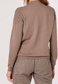 Brauner Pullover mit geripptem Saum und strukturiertem Strickmuster an den Schultern, kombiniert mit Hahnentritt-Musterhose in neutralen Tönen.