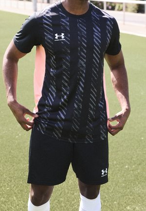 Sportowiec ubrany w czarną sportową odzież marki Under Armour z koszulką i szortami w wzory, stojący na zielonym boisku z sztuczną murawą.