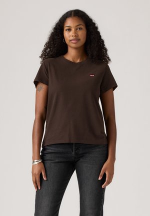 Femme aux cheveux bouclés portant un t-shirt Levi's marron foncé et un jean noir, debout devant un fond clair uni.
