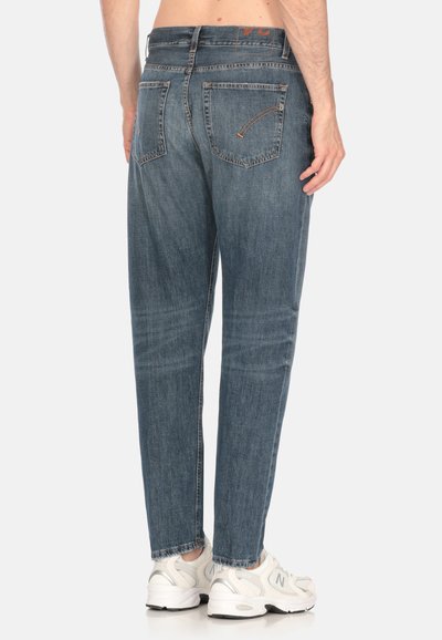 Jeans in denim blu sbiadito con una vestibilità rilassata, dotati di due tasche posteriori e dettagli di cucitura arancioni. Indossati con scarpe da ginnastica bianche.