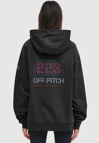 Ciemnoszara bluza z kapturem o luźnym kroju. Posiada duży nadruk graficzny na plecach z napisem "223 OFF PITCH" oraz szczegółami dotyczącymi szerokości i długości geograficznej.
