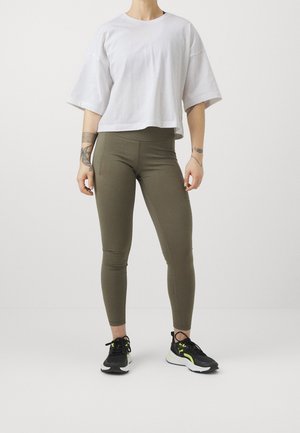 Person, die ein weißes, übergroßes Kurzarmhemd, olivgrüne, hoch taillierte Leggings und schwarze sowie neongrüne Turnschuhe trägt, steht vor einem einfarbigen Hintergrund.