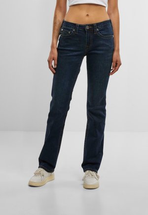 A person stands wearing a fitted white crop top and dark blue jeans, paired with white sneakers, against a plain light background.