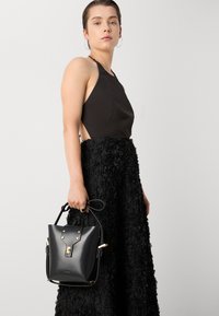 Sac à main en cuir noir avec un haut arrondi, des accents en métal doré, et de courtes poignées. Porté avec une robe noire texturée jusqu'au sol.