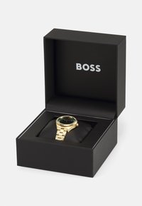 Montre en or avec un cadran vert et un bracelet doré, exposée sur un coussin noir à l'intérieur d'une boîte noire avec le nom de la marque "BOSS" en argent.