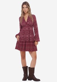 Rood patroonjurk met lange mouwen, een v-hals en een gerimpelde zoom. Gecombineerd met bruine enkelhoge laarzen. Bevat een ingewikkeld paisleypatroon.