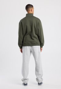Sudadera verde oliva con cuello alto, combinada con pantalones de chándal gris claro. La tela parece suave, con un corte holgado y mangas ajustadas.