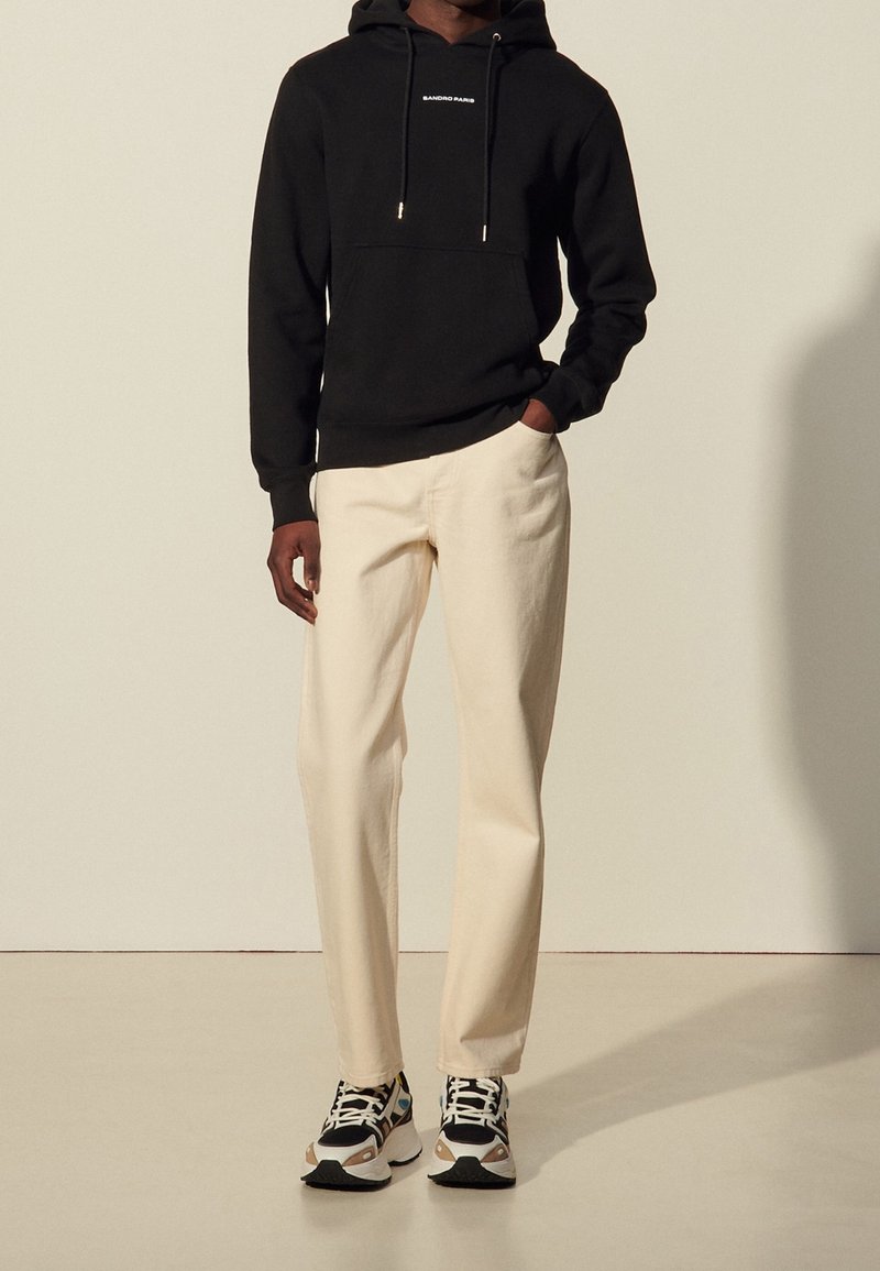 Felpa nera con cappuccio e lacci, pantaloni color crema e sneakers con motivo. Design semplice, materiale in cotone e colori a contrasto.