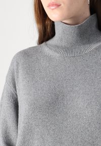 Pull en col roulé gris fabriqué à partir d'un tissu doux et texturé. Il présente un col côtelé et une coupe décontractée avec des épaules tombantes.