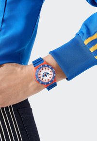 Eri värviline käekell valge näo, oranži äärise, sinise rihma ja suurte numbritega. Silmapaistev spordilik disain ja selge nähtavus.