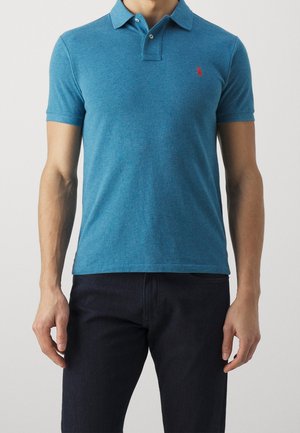 Hombre con una camisa polo azul ajustada de manga corta con cuello y pantalones oscuros sobre un fondo liso.