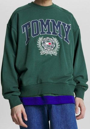 Personne portant un sweat-shirt vert avec "TOMMY" et un blason étiqueté au centre, superposé à une chemise violette et un jean bleu.