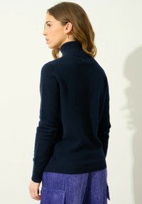 Maglione a collo alto blu navy in tessuto morbido, design aderente, texture a coste, abbinato a pantaloni in corduroy viola con texture, dettagli minimi.