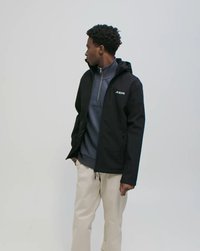 Mladić u crnoj jakni s kapuljačom preko tamnoplavog pulovera i bež hlača, gleda lijevo na jednostavnoj svijetloj pozadini.