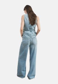 Mono de mezclilla en azul claro, con un cuerpo ajustado, pantalones de pierna ancha y una cintura ceñida con cinturón. La vista trasera muestra dos bolsillos en la parte posterior.