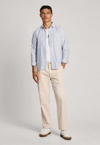 Camisa de manga larga a rayas azul claro y blanca sobre una camiseta blanca, combinada con pantalones beige y zapatillas blancas con suelas marrones.