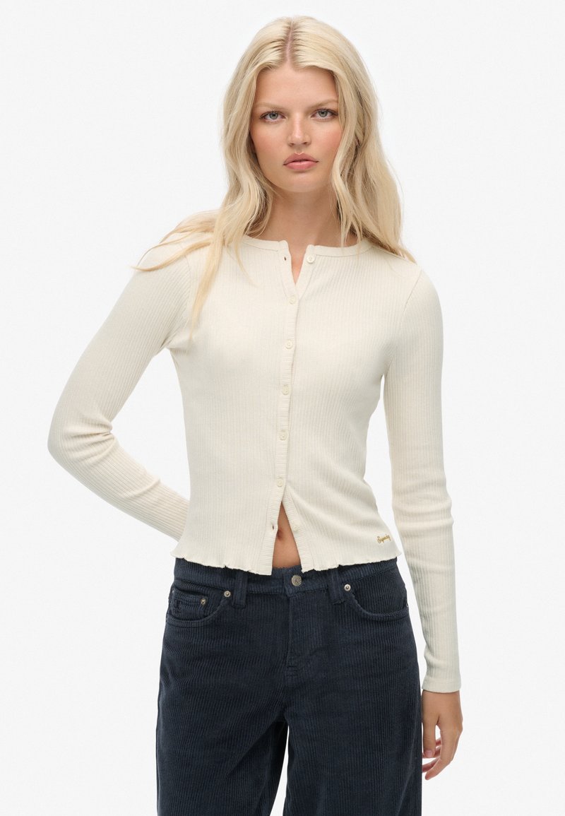 Kremowy, prążkowany kardigan z długim rękawem, okrągłym dekoltem, frontowymi guzikami i lekko skróconym, postrzępionym dołem. W połączeniu z ciemnymi spodniami z kordurowej tkaniny.