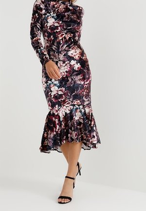 Femme portant une robe à manches longues, longueur genou, fleurie avec un ourlet évasé, associée à des sandales à talons hauts noires avec bride à la cheville.