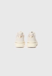Light beige sneakers featuring a smooth upper, mesh accents, and a textured sole. Decorative gold hardware near the heel.