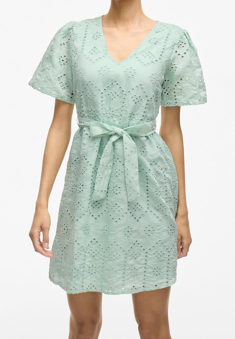 Robe broderie anglaise vert clair avec manches courtes bouffantes, col en V et ceinture nouée à la taille.