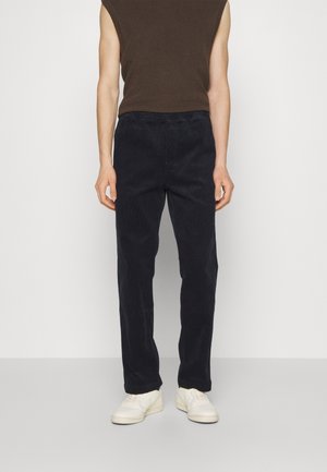 Uomo che indossa pantaloni blu scuro, top marrone senza maniche e sneakers bianche, in piedi davanti a uno sfondo chiaro e uniforme.
