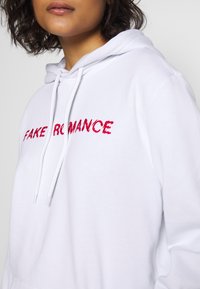 Vit huvtröja med "FAKE ROMANCE" broderat i röd färg. Dragsko i huvan och mjuk textur. Synlig fickdetalj på framsidan.