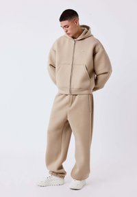 Beige hoodie met ritssluiting en twee voorzakken, gecombineerd met bijpassende joggingbroek. Voorzien van elastische manchetten en een comfortabele pasvorm. Witte sneakers.
