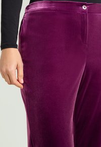 Pantalon en velours de couleur violet profond avec une texture lisse. La taille est dotée d'une fermeture à bouton, avec une main reposant délicatement sur le côté.