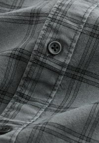 Tessuto a quadri grigio con strisce nere, dotato di un bottone nero e cuciture dettagliate. Materiale testurizzato mostrato da vicino.