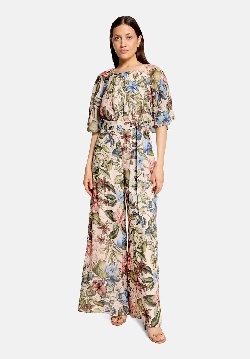Femme en combinaison pantalon large à motif floral, manches bouffantes et ceinture en tissu, debout devant un fond clair uni.